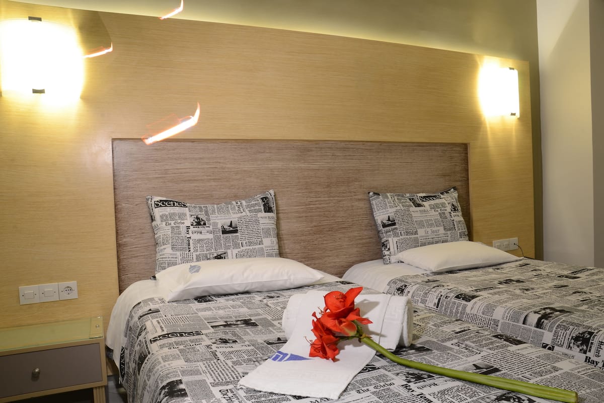 A hotel room with a wooden headboard, two beds with newspaper-themed bedding, a white pillow, and a red rose on a tissue.