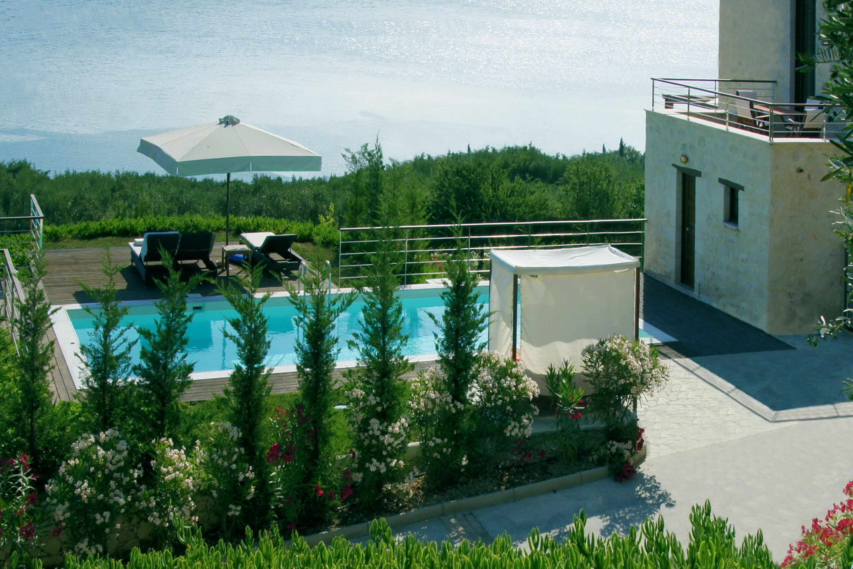 A pool area with lounge chairs and an umbrella, surrounded by greenery and overlooking a body of water.