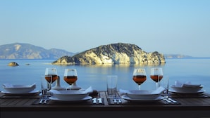 A table set for two with wine glasses and plates, overlooking a calm sea and a rocky island.