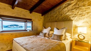A bedroom with a stone wall, wooden ceiling beams, a large bed with pillows, a bedside table with a lamp, and a window with a view of the sea.