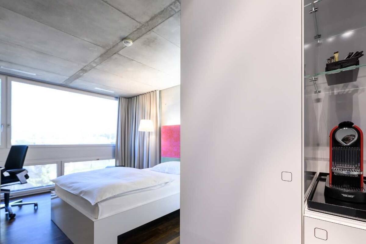 A modern hotel room with a bed, a desk with a chair, a coffee machine, and a large window with curtains.