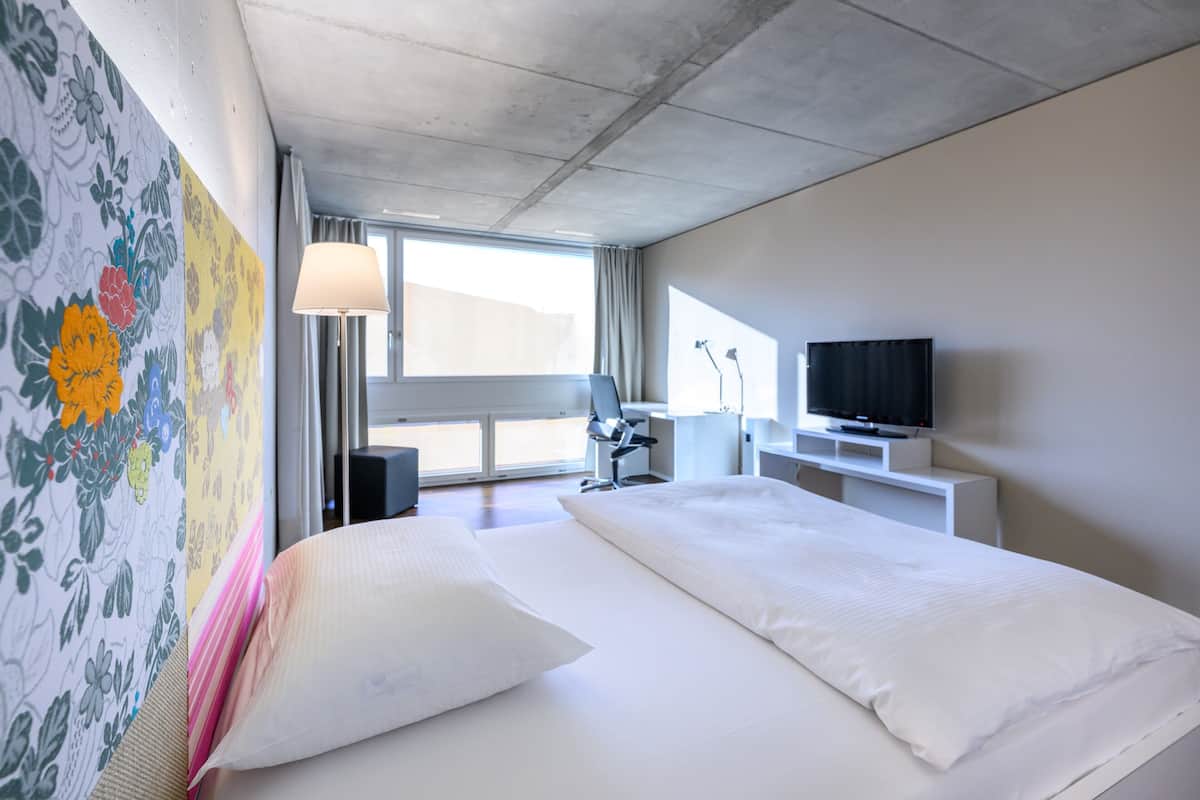 A hotel room with a large bed, a desk, a chair, a TV, and a window with curtains.