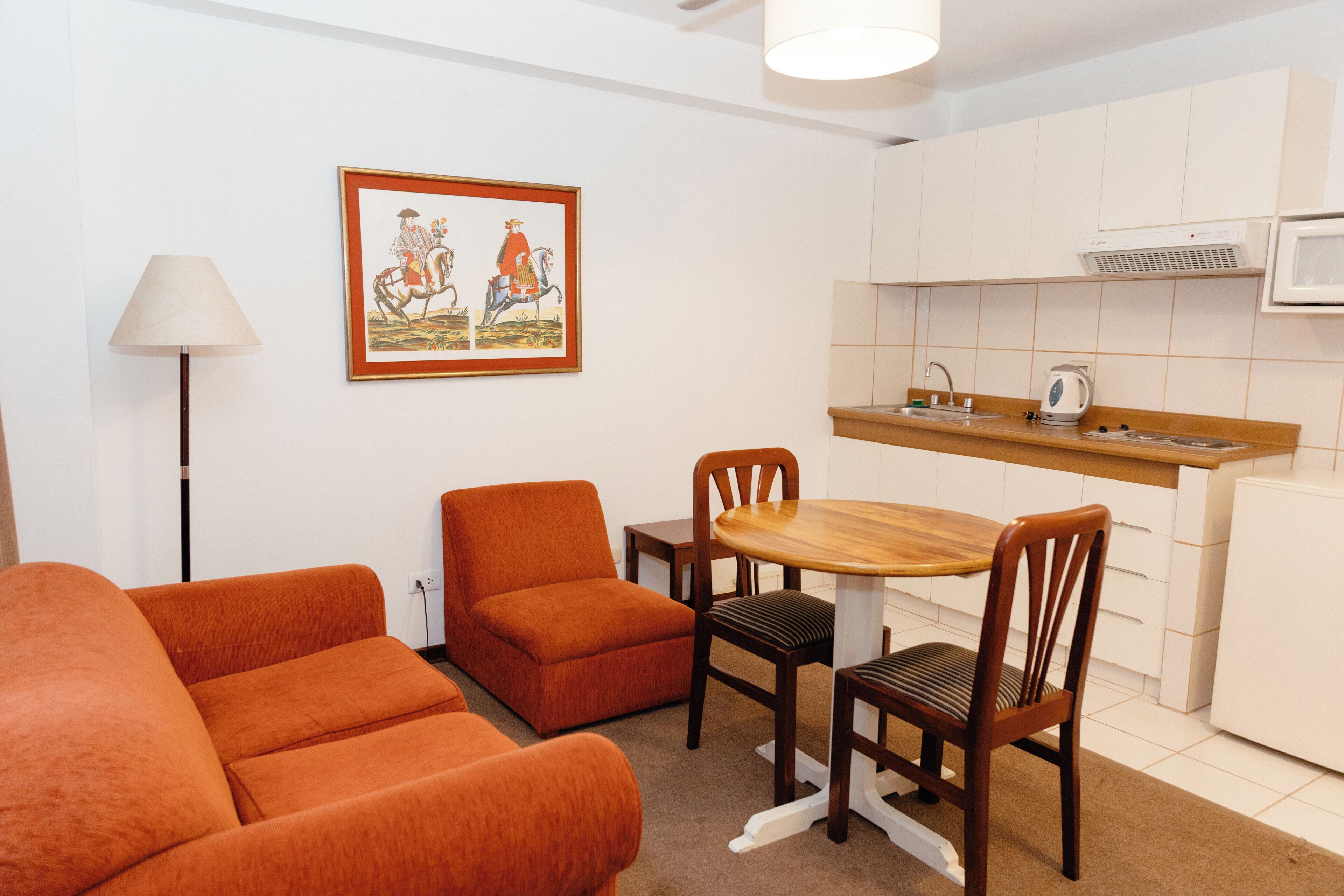 En kompakt opholdsstue med sofa, spisebord med stole, et tekøkken med mikrobølgeovn og en lampe.