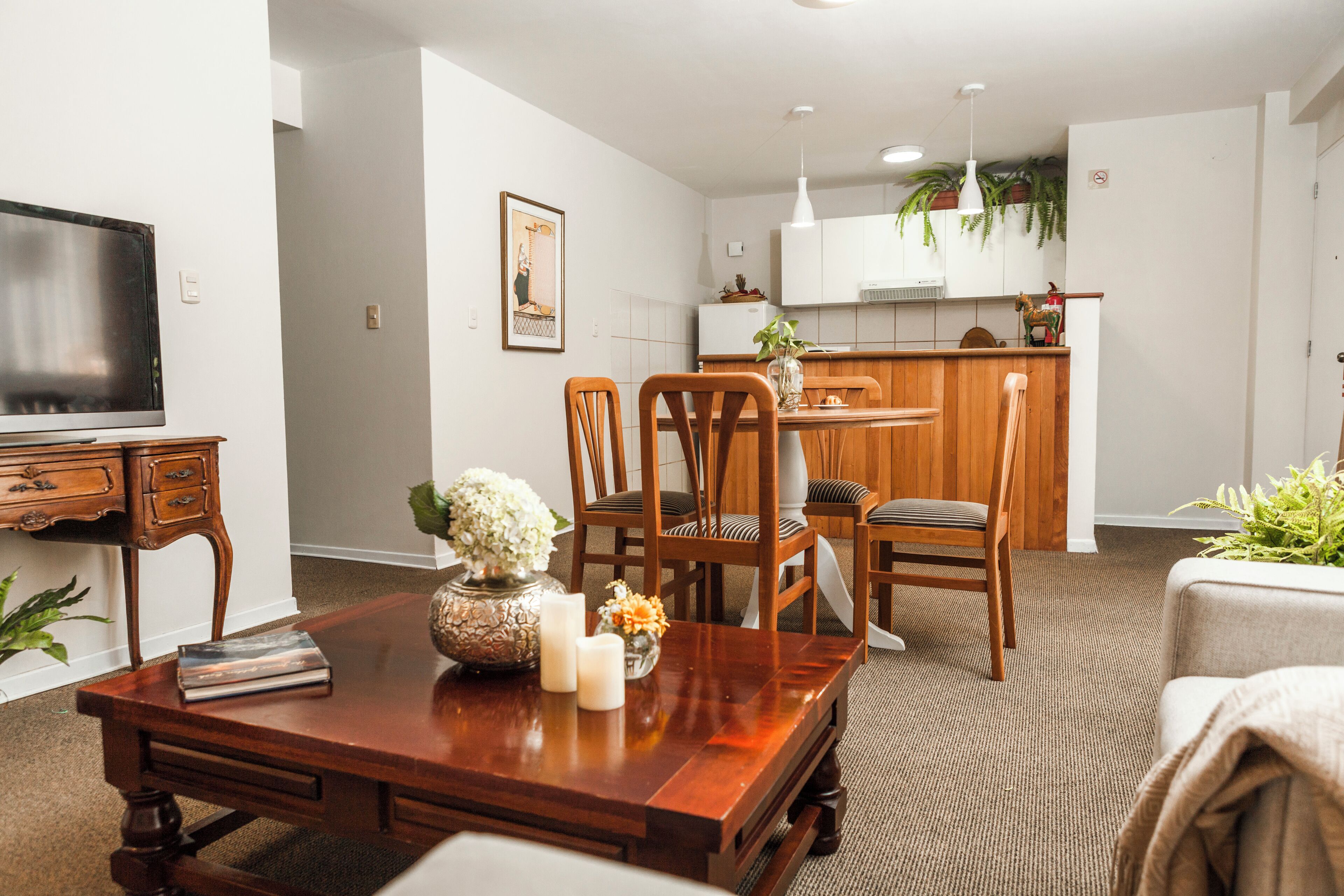 Una sala moderna con una mesa de comedor de madera, dos sillas, un televisor y una zona de cocina al fondo.