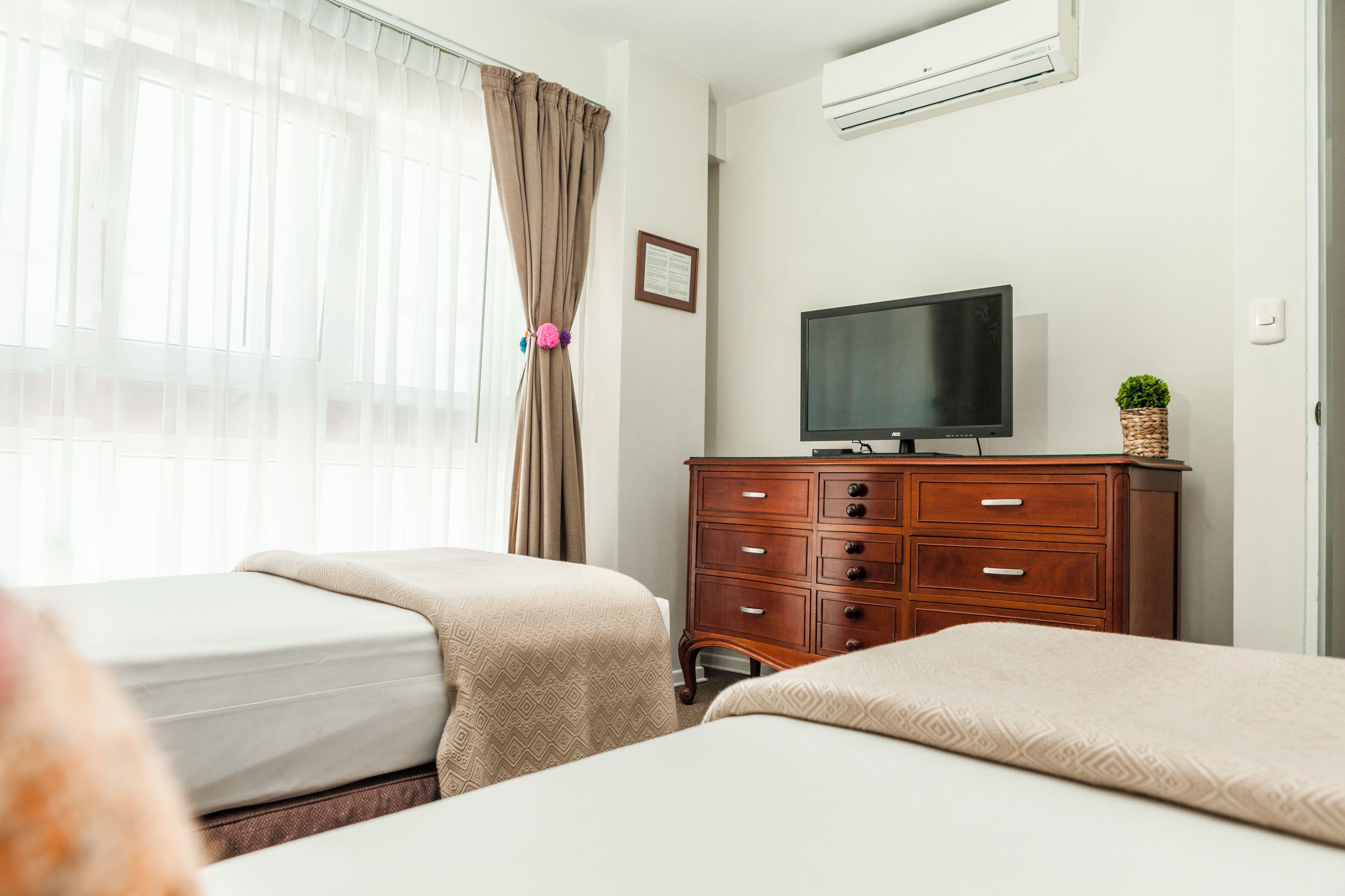 A hotel room with two beds, a wooden dresser, a television, and a window with sheer curtains.