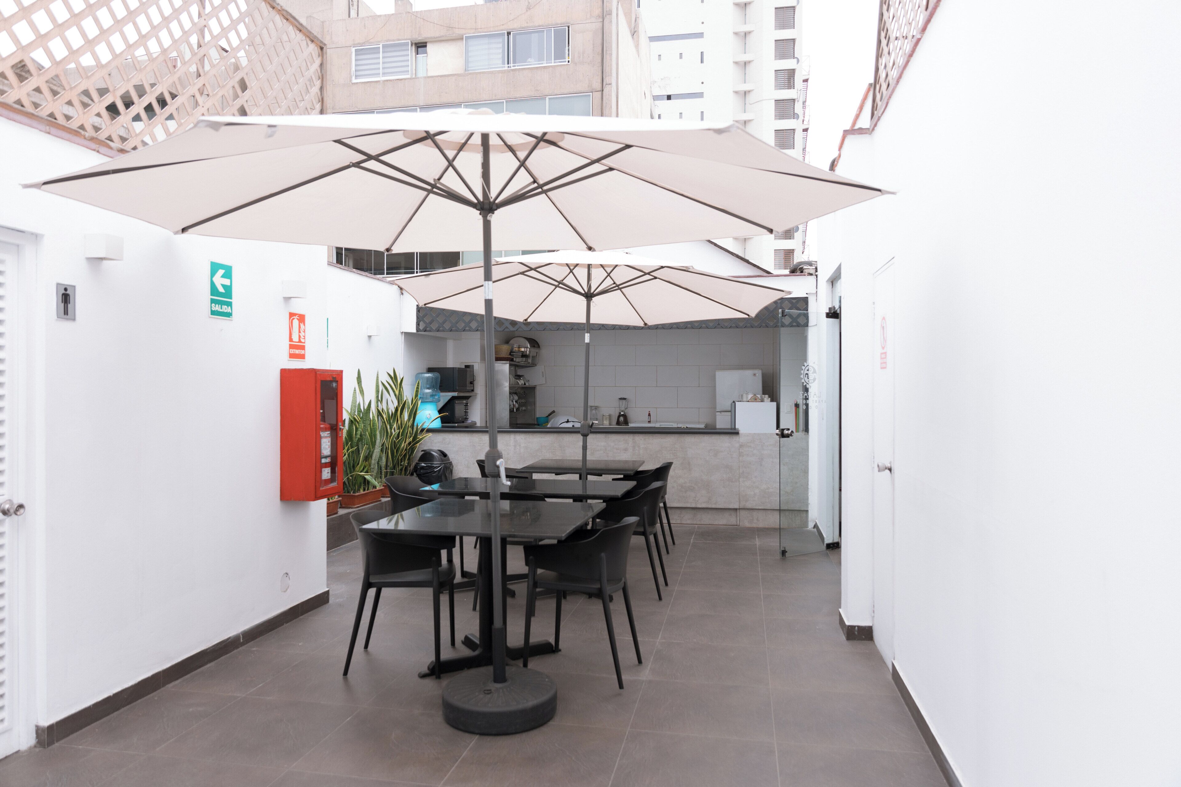 Un área de comedor con una mesa, sillas, un paraguas, un extintor y una cocina al fondo.
