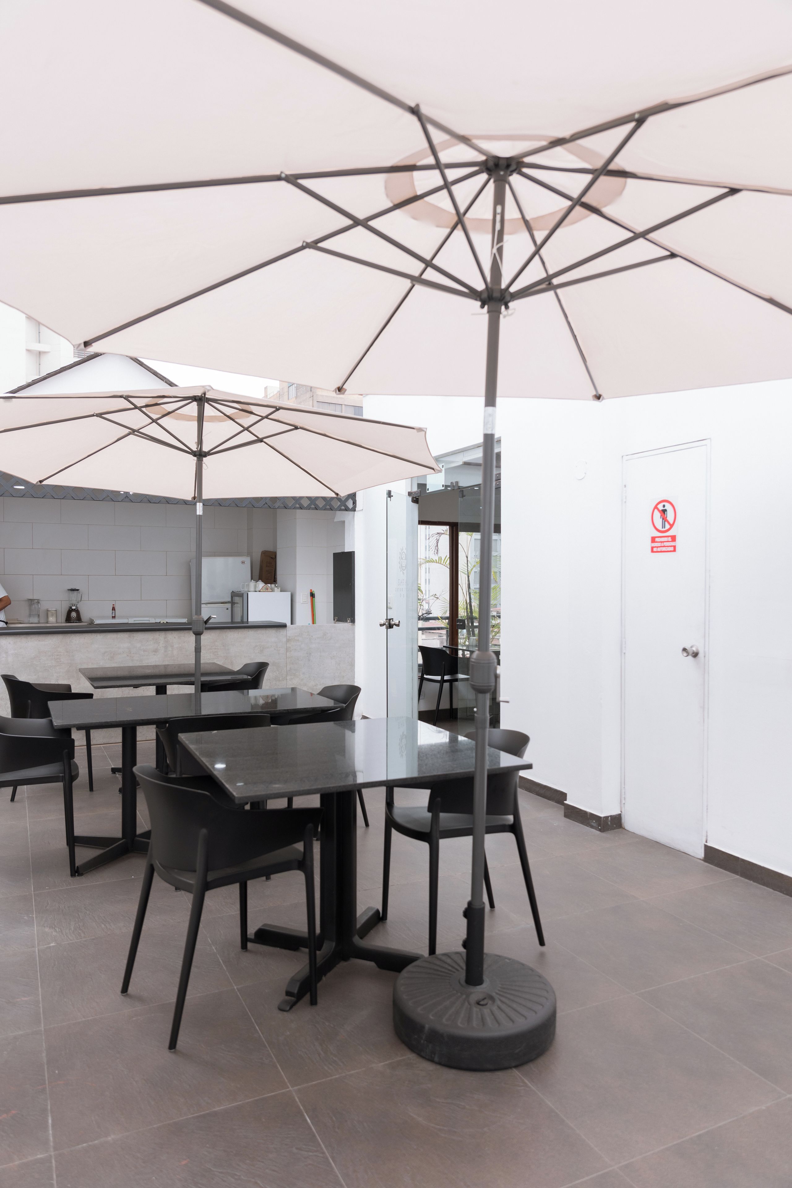 An outdoor seating area with tables and chairs under umbrellas, a kitchen area in the background, and a door with a no-smoking sign.