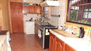 A kitchen with wooden cabinets, a stove, a refrigerator, and a window with a view of greenery outside. - Hostal Emalafquen (Pucon)