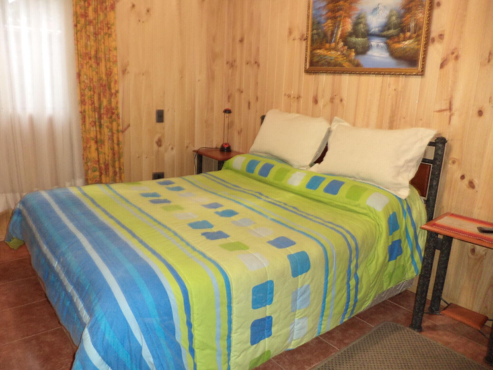 Un dormitorio con pared revestida de madera, una cama con colcha a rayas, un cuadro de un paisaje y una mesita de noche con lámpara.