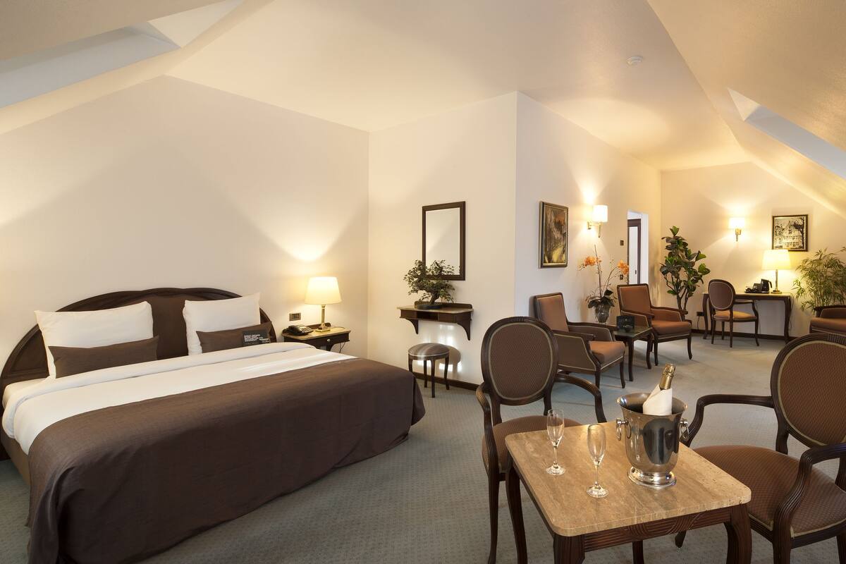 A hotel room with a bed, a TV, a dining table with chairs, and a small seating area with a sofa and chairs.