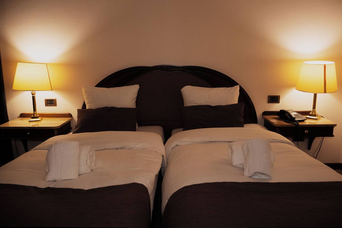 A hotel room with two beds, each with a pillow and a blanket. There are two bedside tables, each with a lamp and a phone.