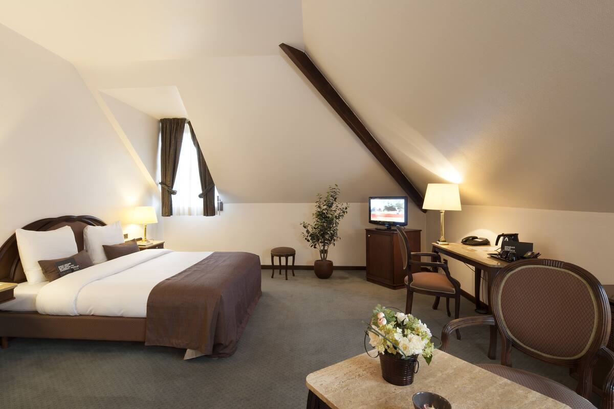 A hotel room with a bed, desk, chair, and a small table with a flower vase.