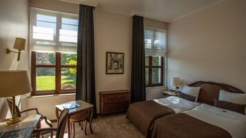 Quarto de hotel com duas camas, uma escrivaninha, uma cadeira, uma mesinha, um abajur e uma janela com vista para áreas verdes.