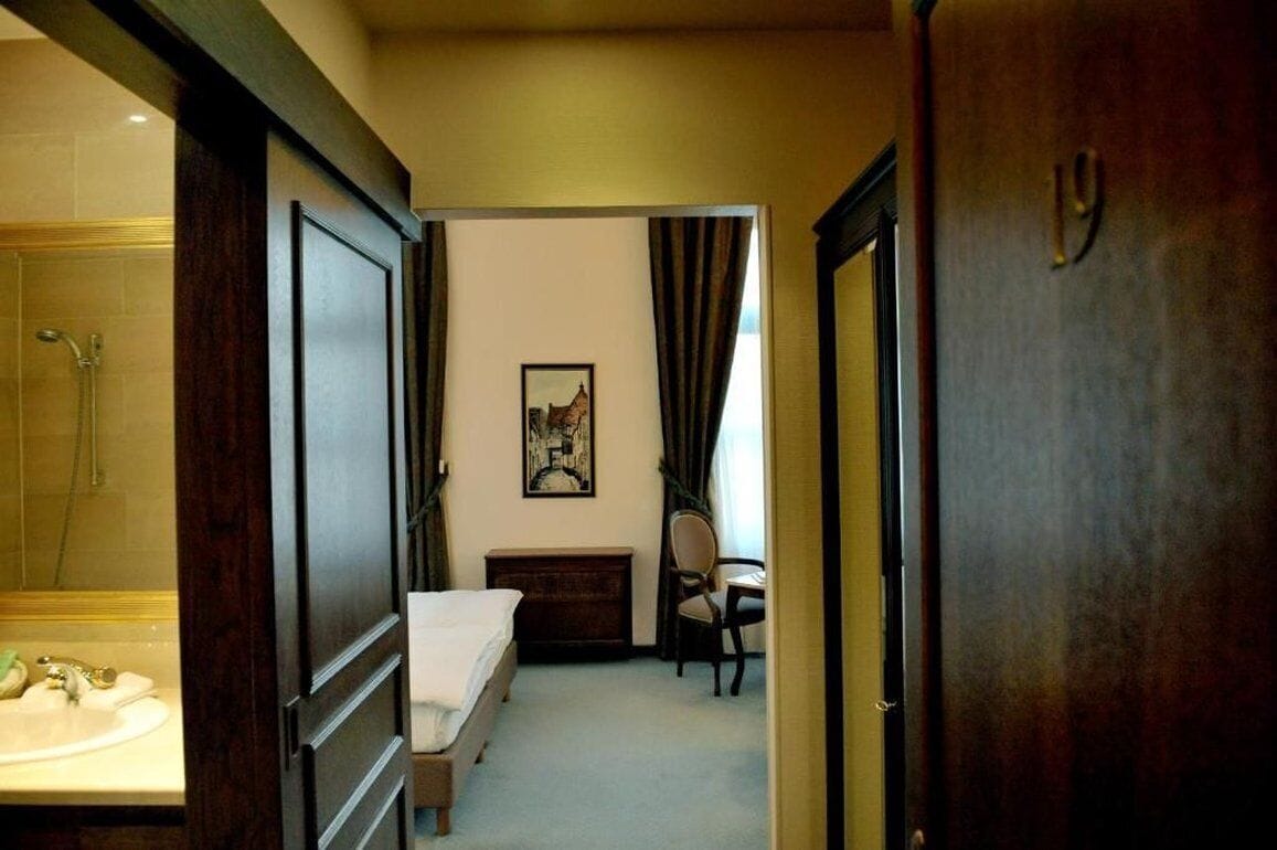 A hotel room with a bed, a desk, a chair, a small table, and a bathroom with a sink and shower.