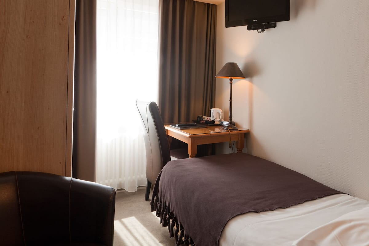 A hotel room with a bed, a desk with a lamp, a chair, and a TV mounted on the wall.