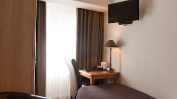 Quarto de hotel com cama, escrivaninha com luminária, cadeira e televisão fixada na parede.