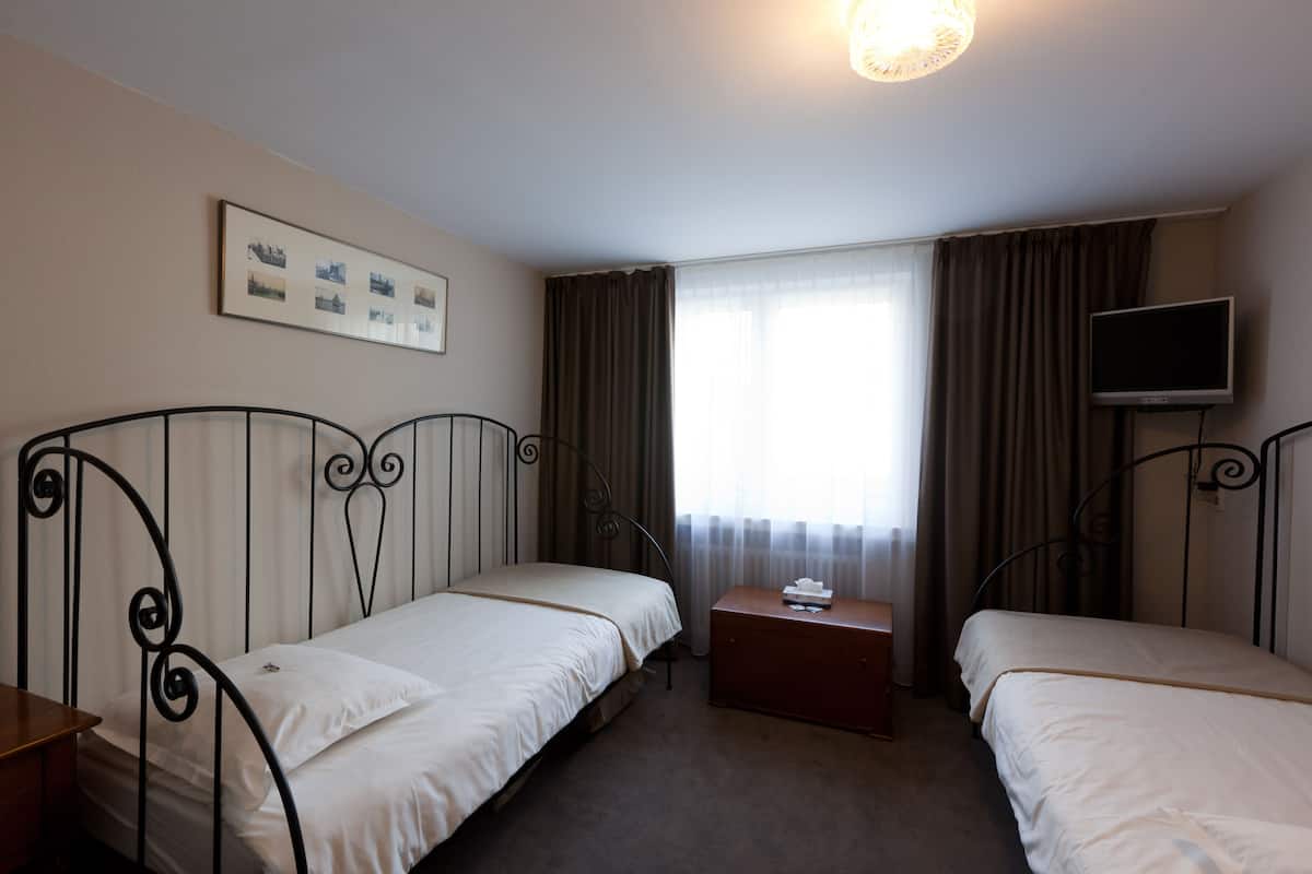 A hotel room with two single beds, a small wooden nightstand, a television on the wall, and a window with curtains.