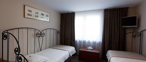 A hotel room with two single beds, a small wooden nightstand, a television on the wall, and a window with curtains.