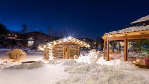 Une cabane en rondins dotée d’une véranda en verre, entourée de neige et illuminée par des lumières.
