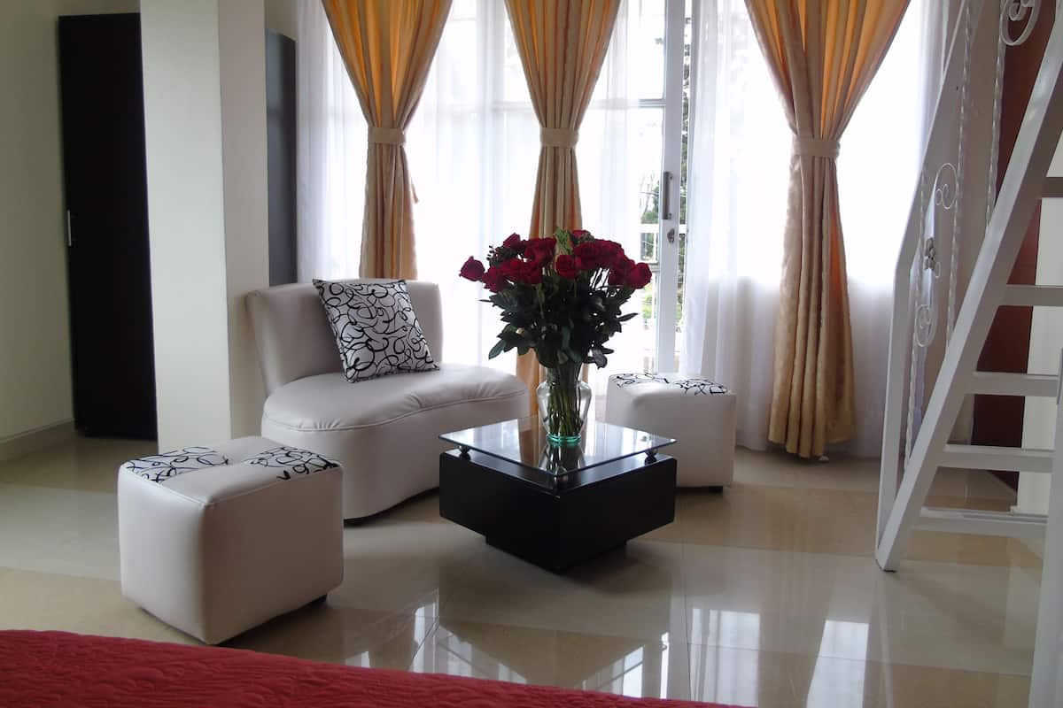 A modern living room with a white sofa, a glass coffee table, and a red rug. There are two white ottomans, a vase with red flowers, and a staircase with a white railing.