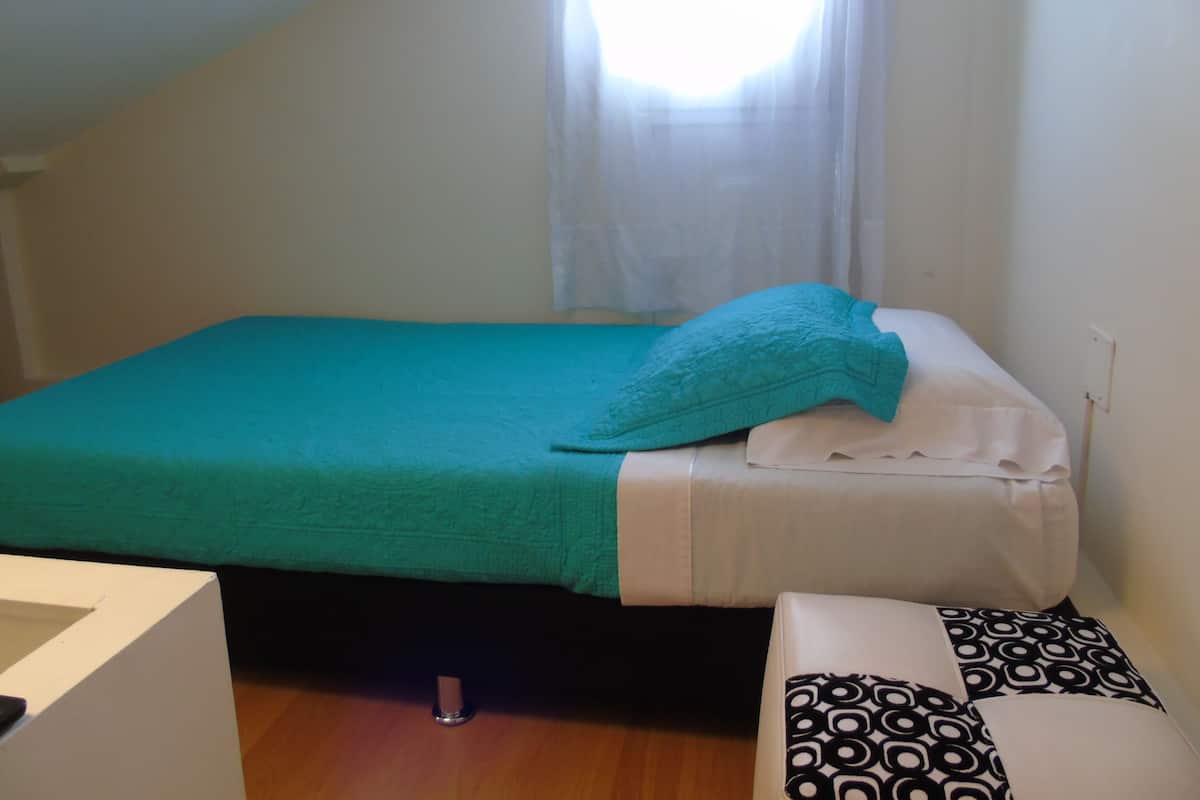 A small, single-bed room with a teal bedspread, white pillows, and a black and white patterned pillow. There is a small bedside table with a lamp, and a window with a white curtain.