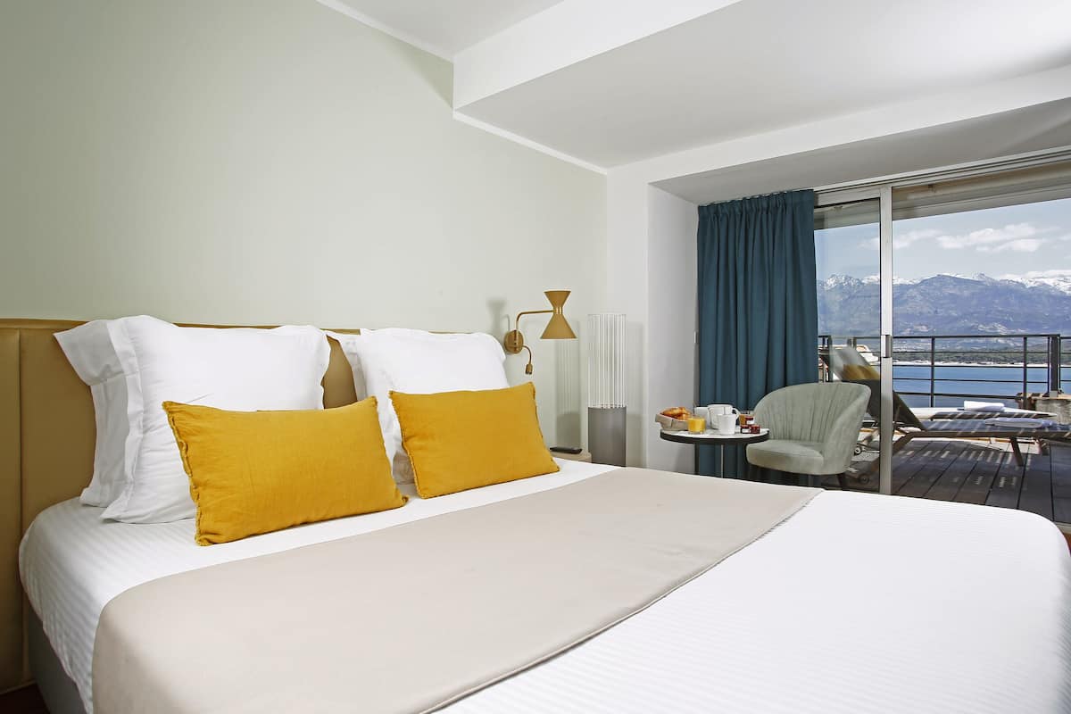 Una habitación de hotel con cama, una silla, una mesa con una taza y vistas a las montañas y al agua.