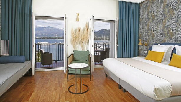 A hotel room with a bed, a sofa, a small table, and a chair. There is a balcony with a view of the sea and mountains. - Hotel Balanea (Calvi)