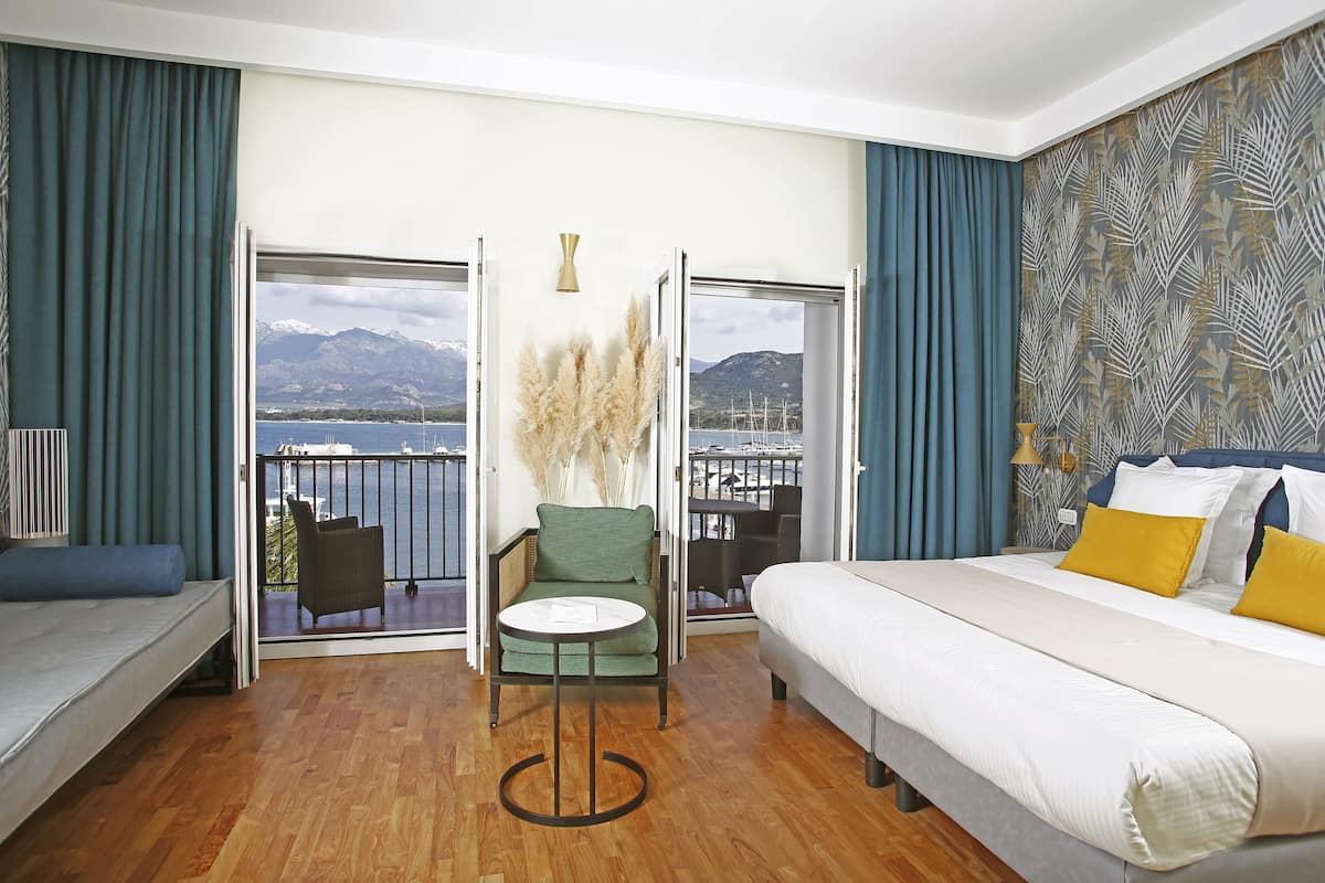 Habitación de hotel con cama, sofá, una mesita y una silla. Tiene un balcón con vistas al mar y a las montañas.