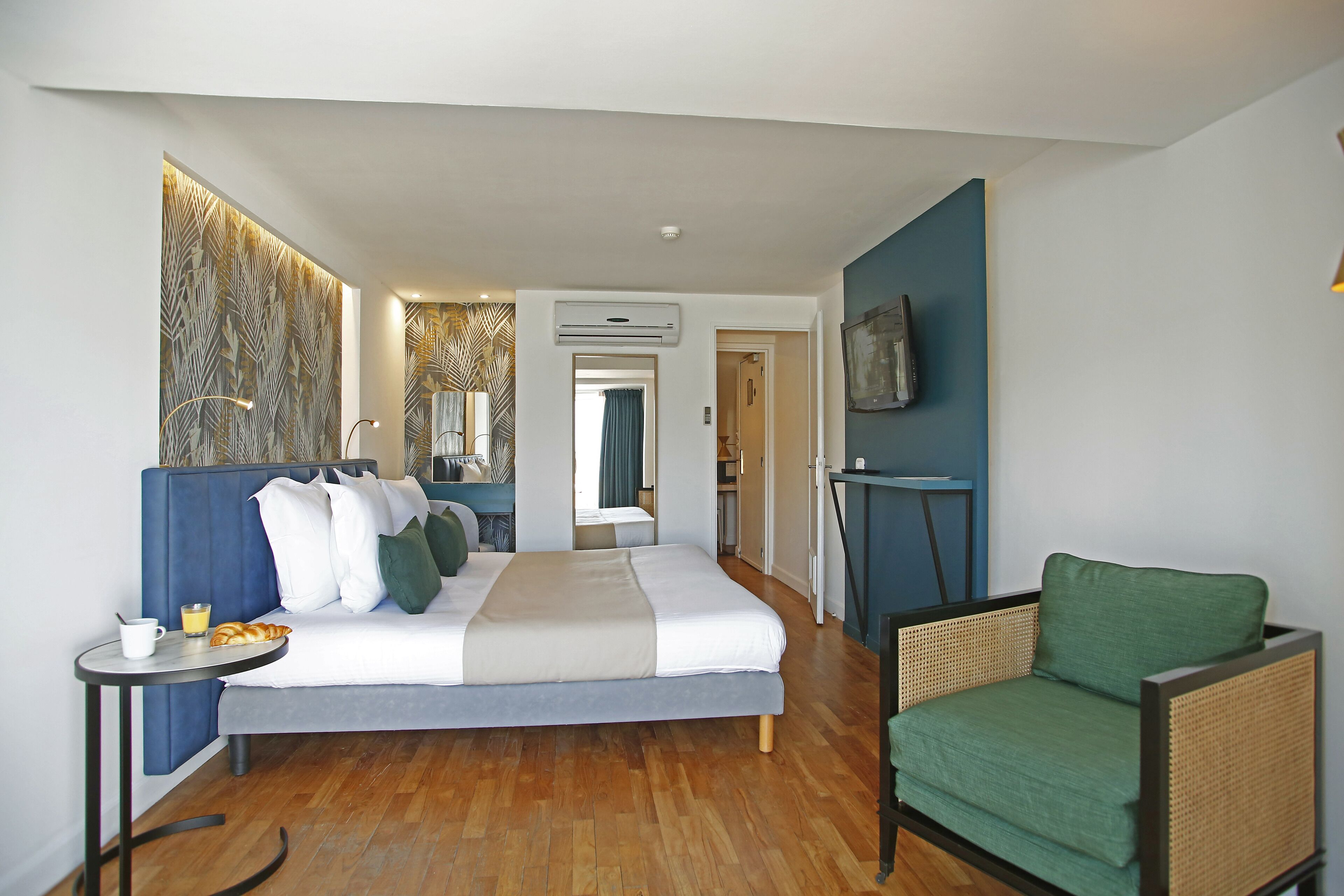 A modern hotel room with a large bed, a green armchair, a small table with a cup and croissant, and a wall-mounted TV.