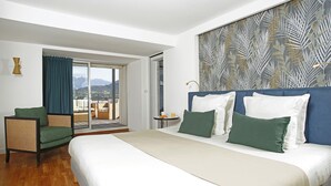 A hotel room with a large bed, a chair, a small table with a cup and a book, and a view of mountains through a sliding door. - Hotel Balanea (Calvi)