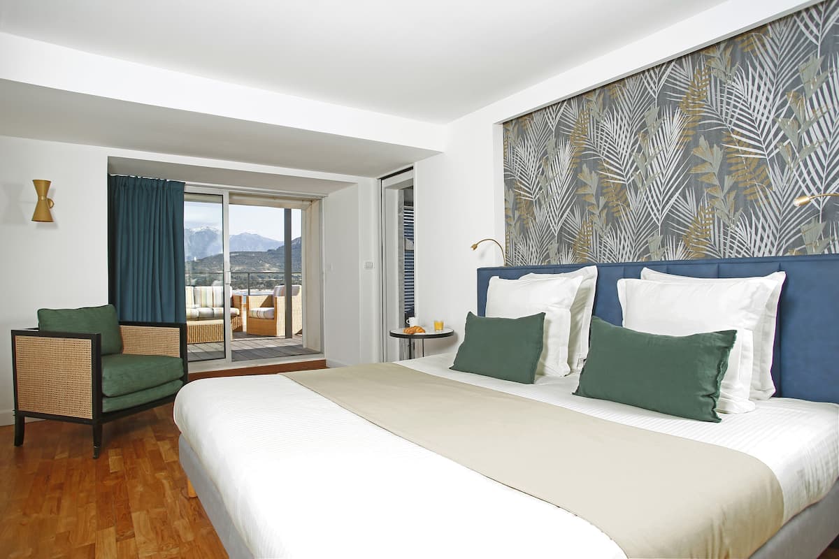 Habitación de hotel con una cama grande, una silla, una mesita con una taza y un libro, y vistas a montañas a través de una puerta corrediza.