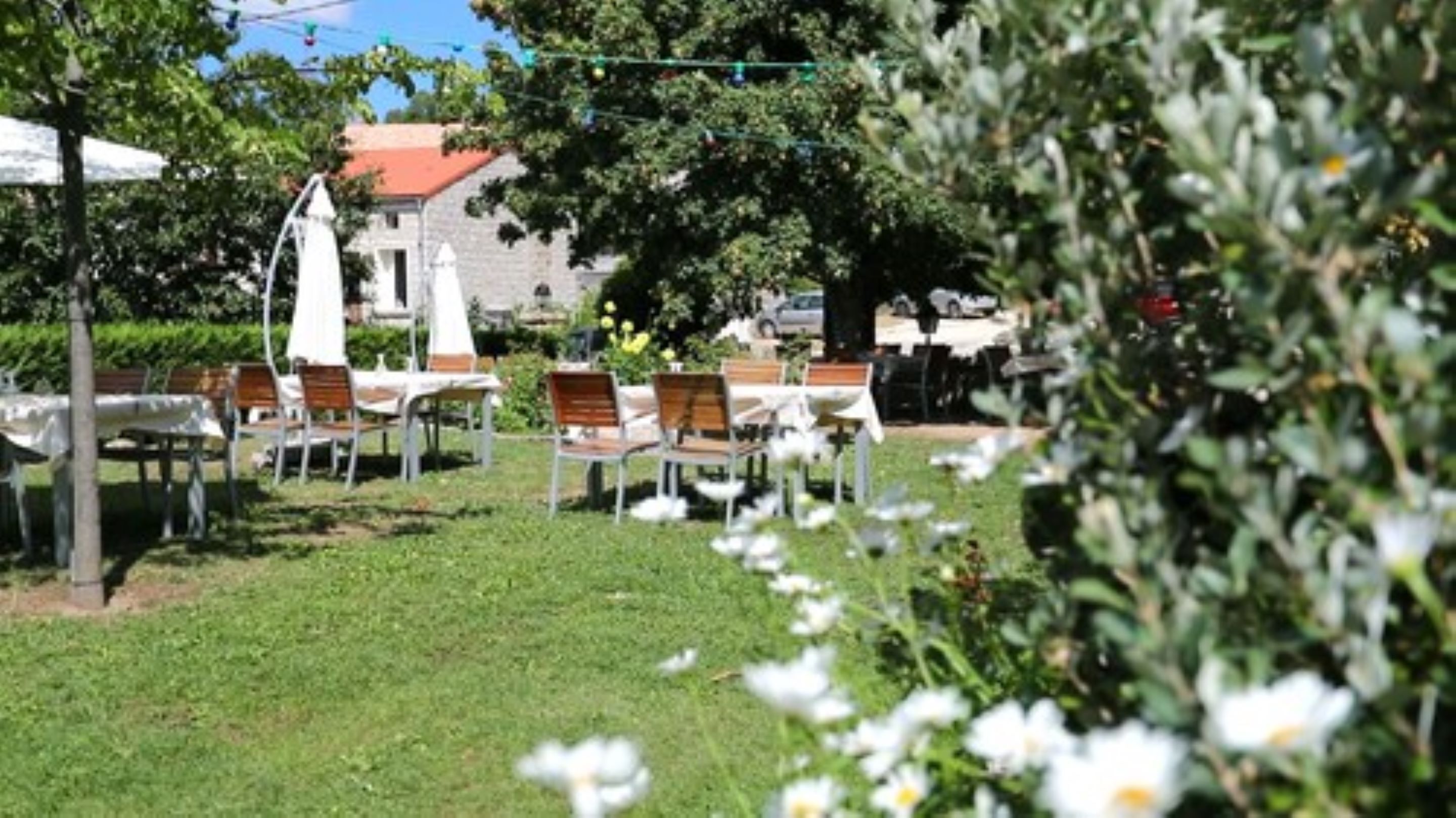 An outdoor seating area with tables and chairs, surrounded by greenery and a building in the background.