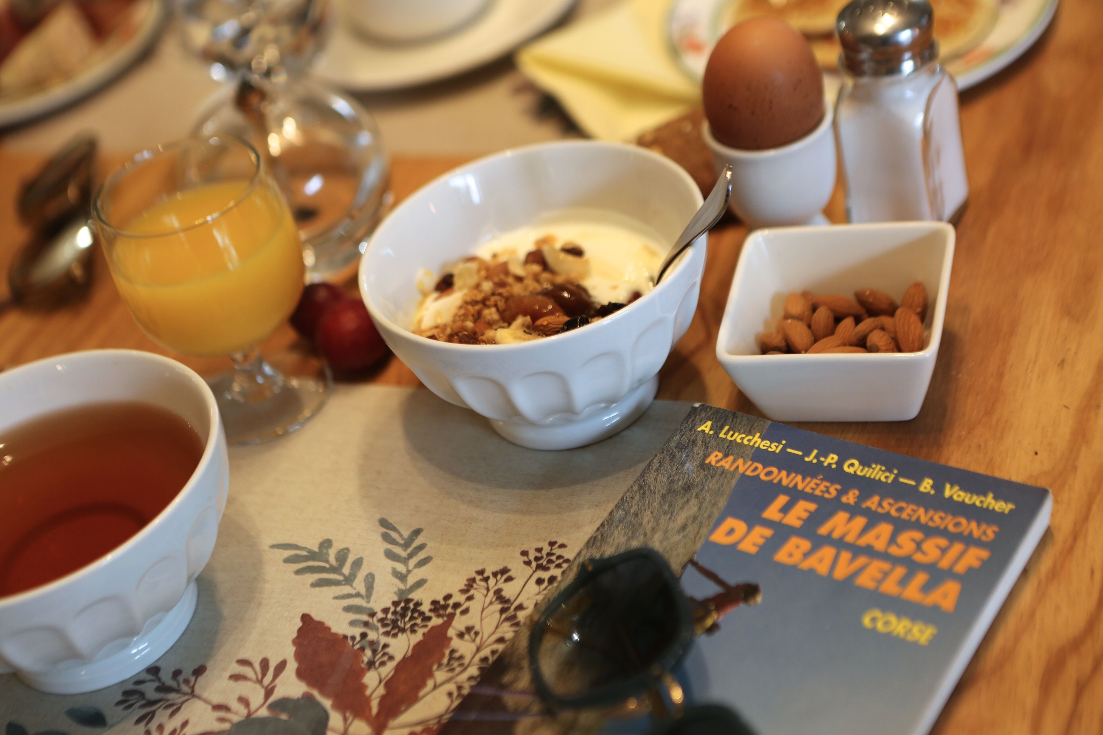 A breakfast table with a bowl of cereal, a glass of orange juice, a cup of tea, and a book titled "Le Massif de Bavella".