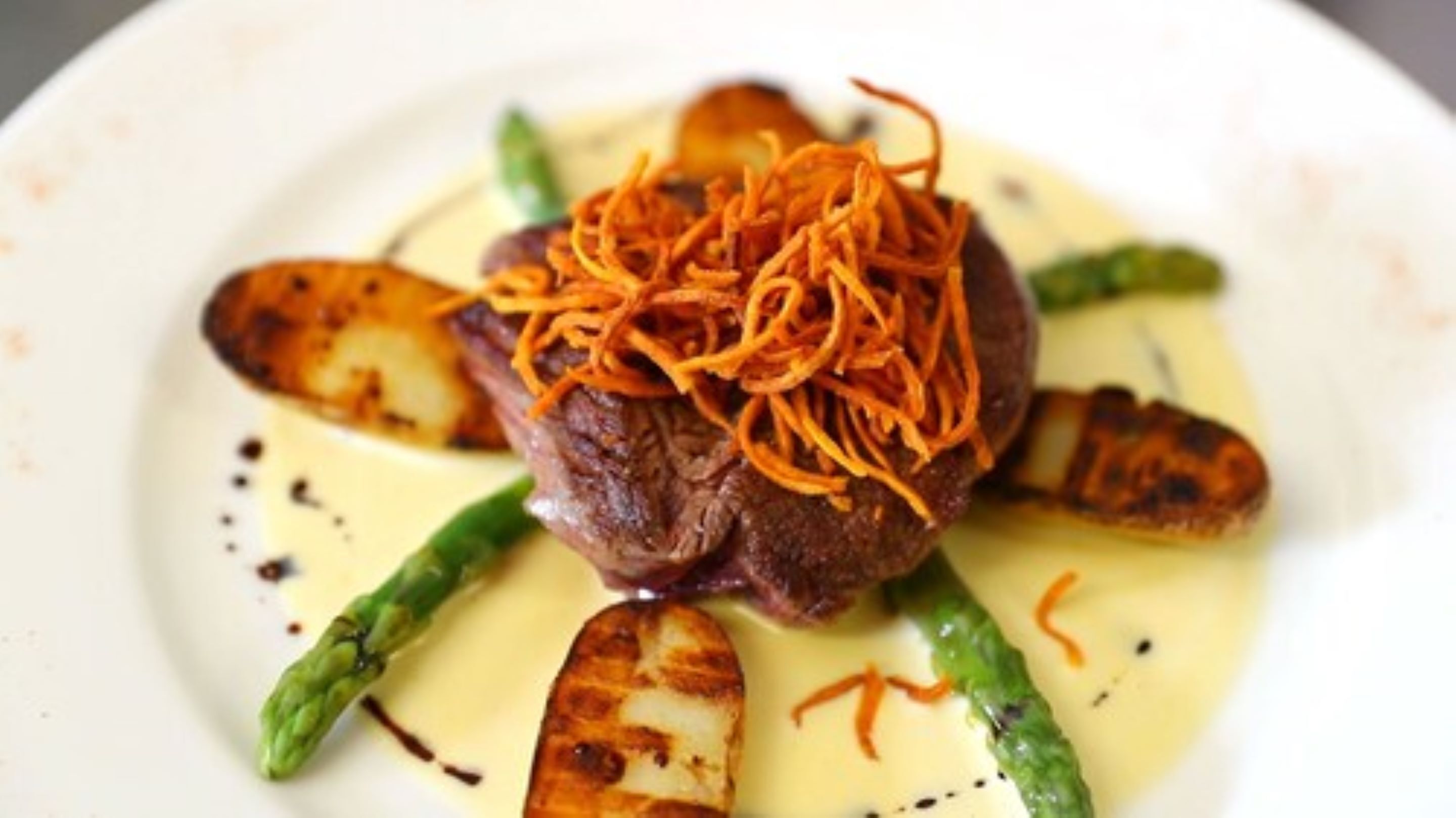 A plated dish with a steak topped with carrot shreds, asparagus, and roasted potatoes.