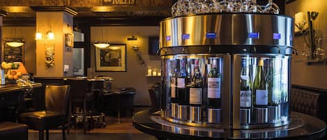 Zona de bar con dispensador de bebidas de acero inoxidable, copas de vino encima y un cartel que indica el camino a los baños.