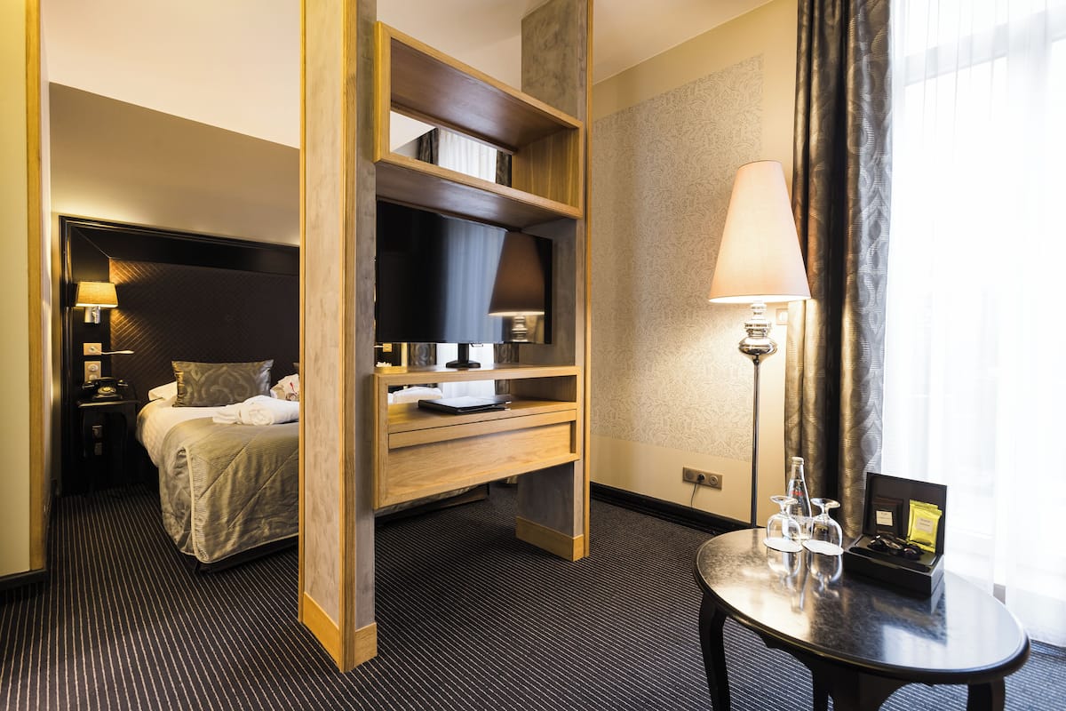 Una habitación de hotel con una cama, un escritorio con lámpara, una mesita con vasos y una botella, y una ventana con cortinas.