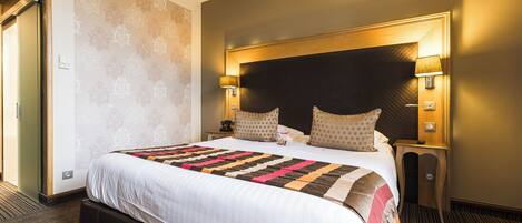 Habitación de hotel con una cama grande, dos lámparas de noche, un cabecero de madera y suelo cubierto de alfombra.