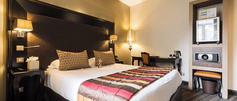 A hotel room with a large bed, two bedside lamps, a desk with a chair, a trash bin, and a refrigerator.