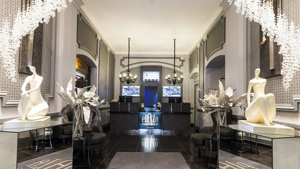 Un hall d’hôtel moderne avec un comptoir d’accueil central, deux grandes sculptures blanches et un écran numérique.