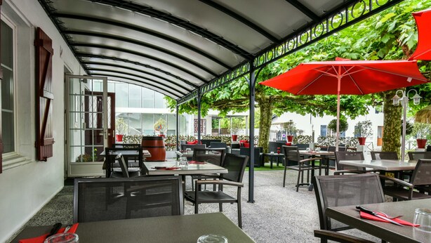 An outdoor dining area with tables and chairs, covered by a curved roof, and surrounded by greenery. - Auberge L'Estanquet (Urt)