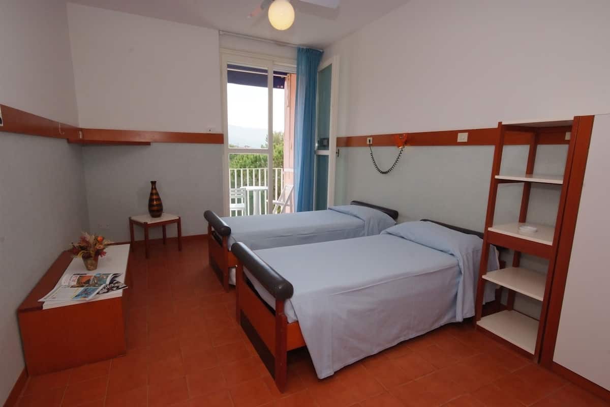 Een kamer met twee eenpersoonsbedden, een klein tafeltje met een vaas en een krant, een houten wandplank en een balkon met een blauw gordijn.