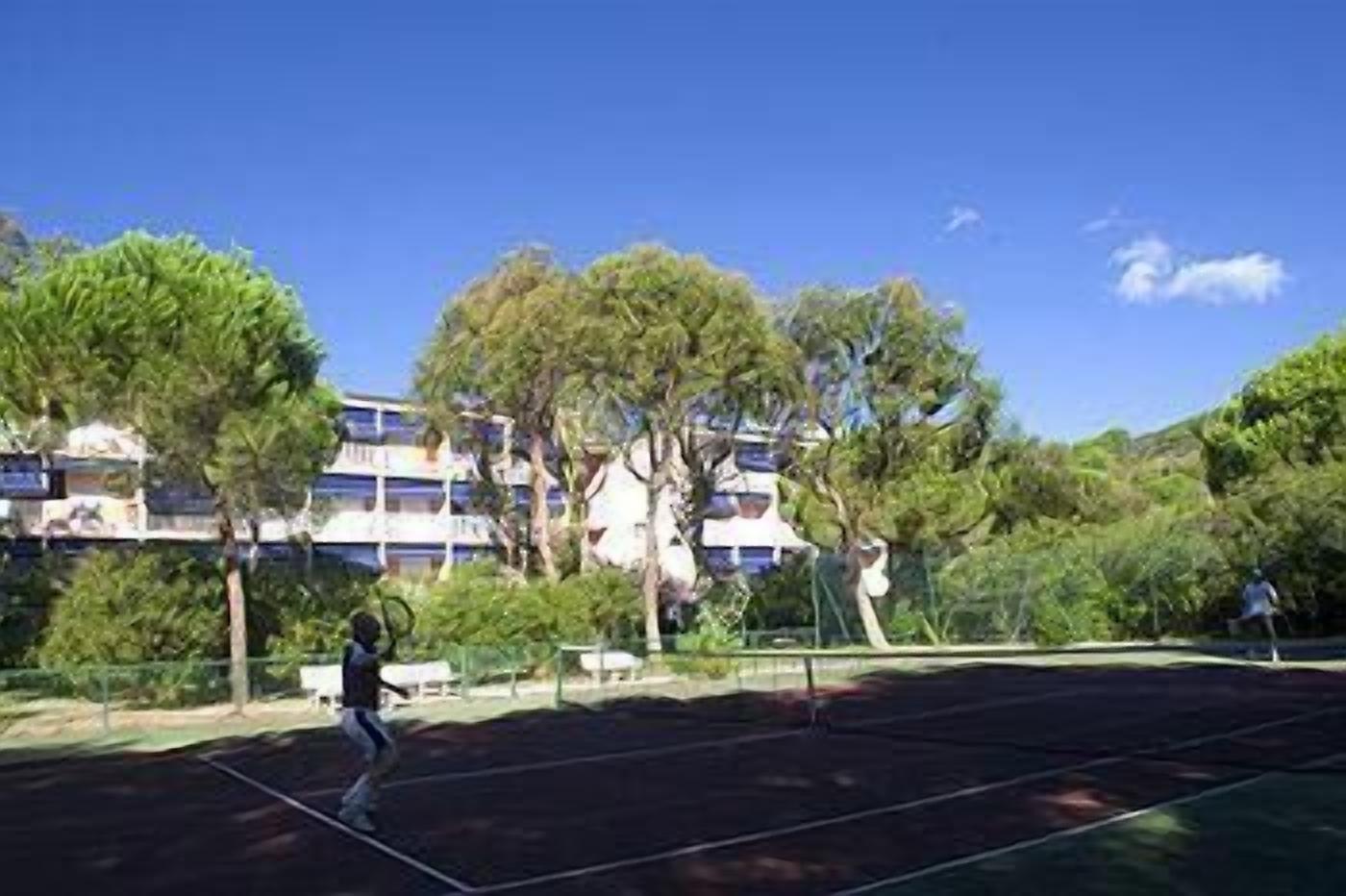 Una cancha de tenis con red, rodeada de árboles y un edificio al fondo.