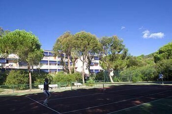 Una cancha de tenis con red, rodeada de árboles y un edificio al fondo.