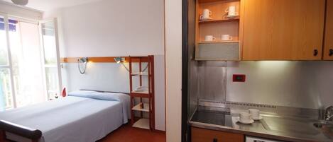 A bedroom with a bed, a wooden nightstand, and a small kitchenette with a sink and cabinets.