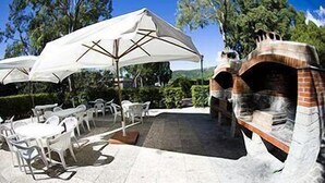 Um pátio externo com cadeiras e mesas de plástico branco, guarda-sóis brancos e um forno de tijolos.