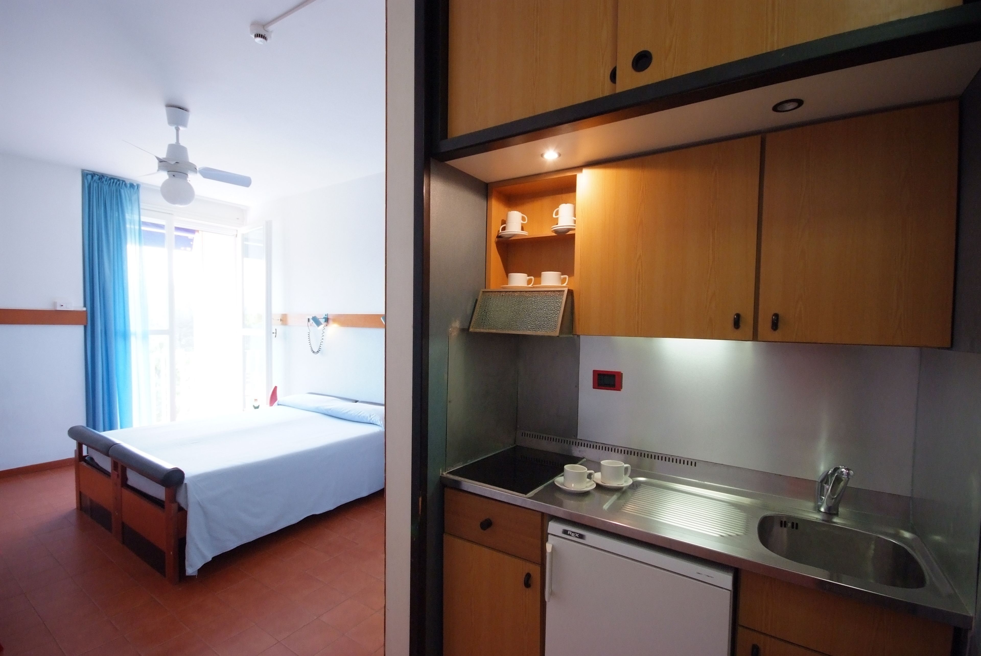 Una habitación con cama, ventana con cortinas, ventilador de techo y kitchenette con pileta y armarios.