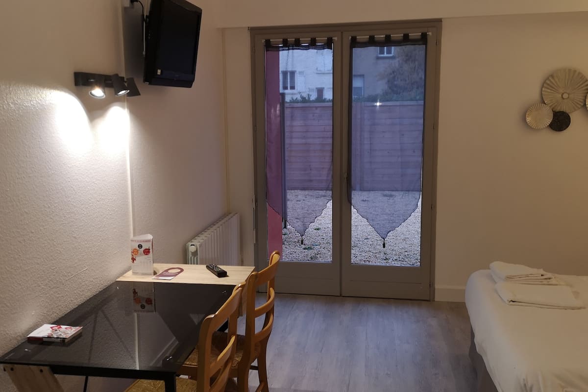 A compact room with a small table, a chair, a sofa, and a sliding glass door leading to an outdoor area.