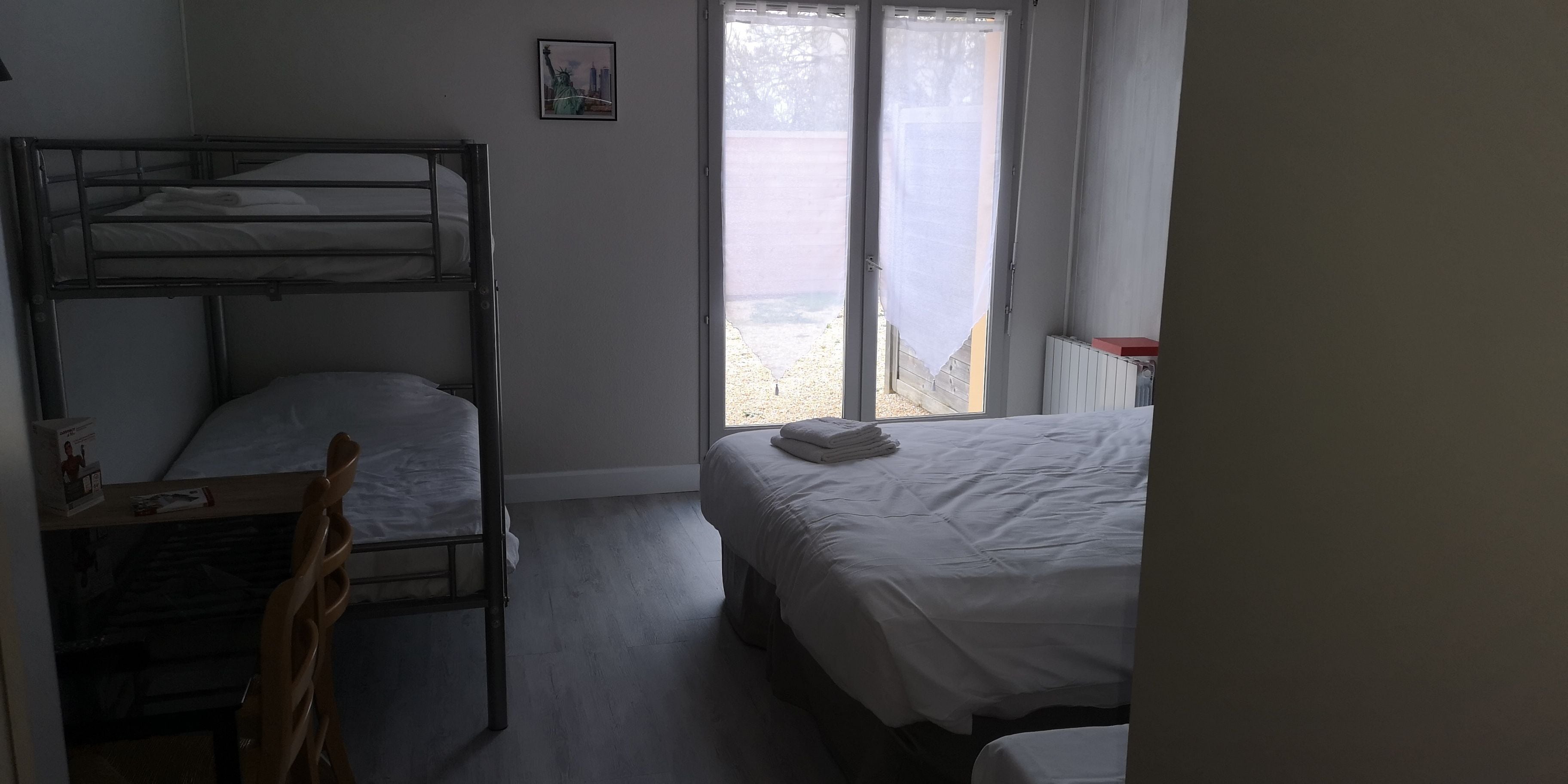 A dormitory room with bunk beds, a window with curtains, and a small bedside table.
