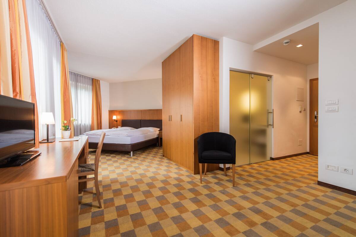 A hotel room with a checkered floor, a bed, a desk with a chair, a TV, and a wardrobe.