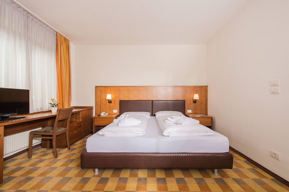 A hotel room with a bed, a desk, a chair, a television, and a nightstand with lamps.